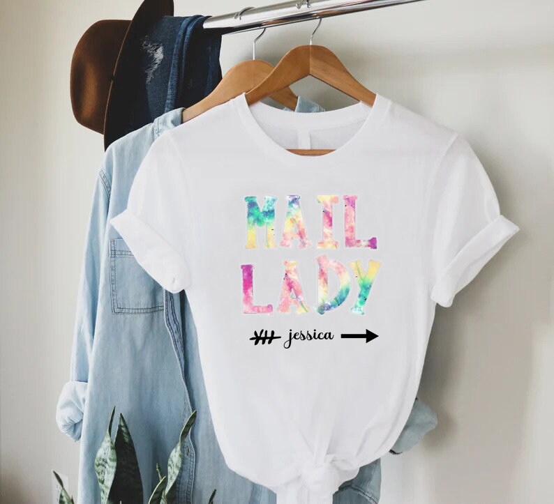 May include: White t-shirt with a tie-dye effect design that says "Mail Lady" with the name "Jessica" written below it.