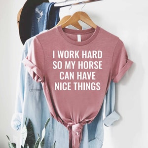 Może przedstawiać: R&oacute;żowy T-shirt z nadrukiem "Pracuję ciężko, żeby m&oacute;j koń miał fajne rzeczy" w kolorze białym.