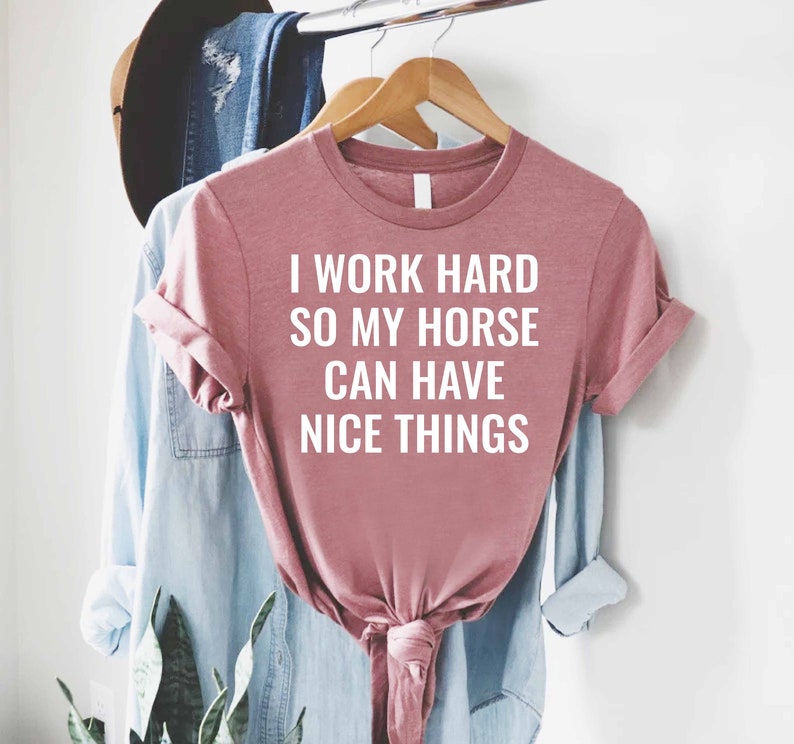 Może przedstawiać: R&oacute;żowy T-shirt z nadrukiem "Pracuję ciężko, żeby m&oacute;j koń miał fajne rzeczy" w kolorze białym.
