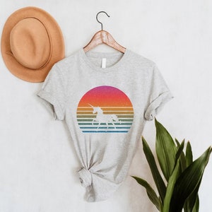 K&ouml;nnte beinhalten: Hellgraues T-Shirt mit einem Retro-Design eines Einhorns in einem farbenfrohen Sonnenuntergang. Das Design umfasst horizontale Streifen in Orange, Gelb, Gr&uuml;n und Blau. Das T-Shirt h&auml;ngt an einem Holzkleiderb&uuml;gel.