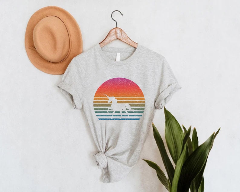 K&ouml;nnte beinhalten: Hellgraues T-Shirt mit einem Retro-Design eines Einhorns in einem farbenfrohen Sonnenuntergang. Das Design umfasst horizontale Streifen in Orange, Gelb, Gr&uuml;n und Blau. Das T-Shirt h&auml;ngt an einem Holzkleiderb&uuml;gel.