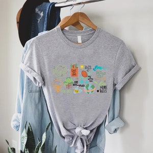Puede incluir: Camiseta gris con un dise&ntilde;o gr&aacute;fico colorido que presenta una variedad de mensajes ambientales, que incluyen "Salvemos nuestros mares", "Este tambi&eacute;n es mi hogar", "No uses pl&aacute;stico" y "M&aacute;s &aacute;rboles".