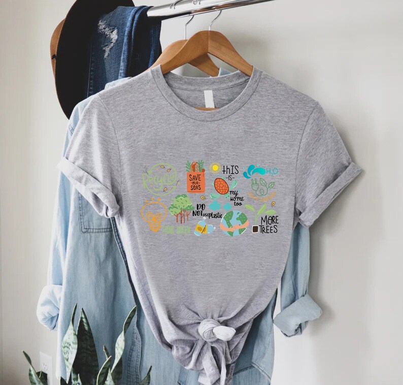 Puede incluir: Camiseta gris con un dise&ntilde;o gr&aacute;fico colorido que presenta una variedad de mensajes ambientales, que incluyen "Salvemos nuestros mares", "Este tambi&eacute;n es mi hogar", "No uses pl&aacute;stico" y "M&aacute;s &aacute;rboles".