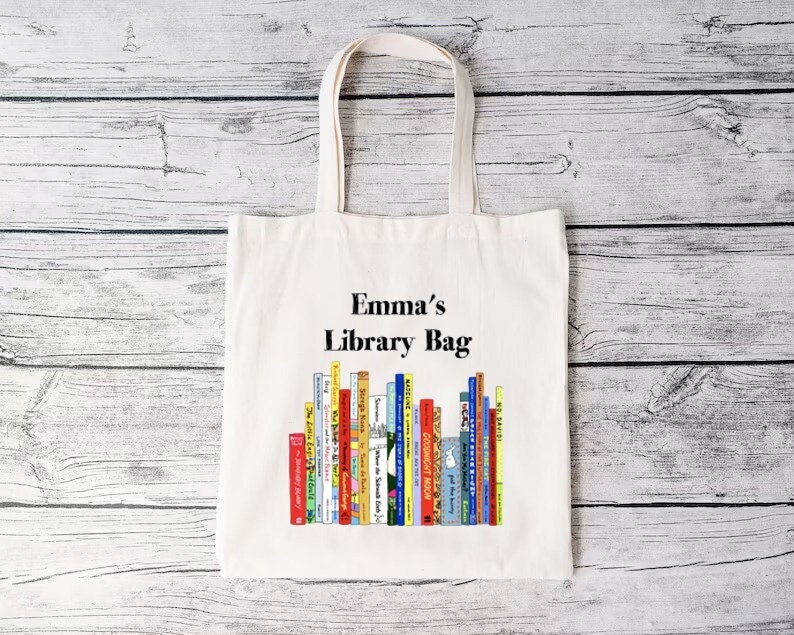 May include: A white canvas tote bag with the text "Emma's Library Bag" above a colorful illustration of a stack of books. The bag has two long handles for carrying and is set against a light wood background.