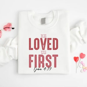 Puede incluir: Sudadera blanca con el texto "He Loved Us First 1 John 4:19" en letras rosas. La sudadera est&aacute; doblada y se muestra sobre un fondo blanco con gafas de sol en forma de coraz&oacute;n rosas y corazones de caramelo.