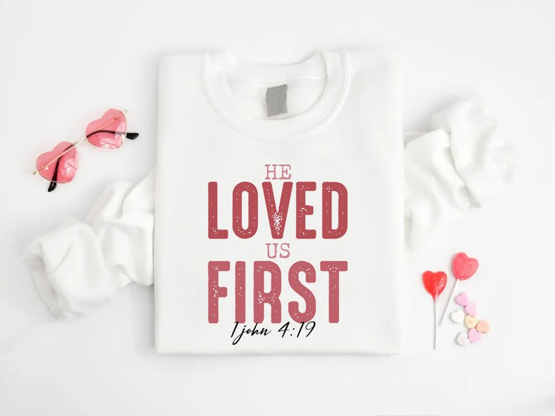 Puede incluir: Sudadera blanca con el texto "He Loved Us First 1 John 4:19" en letras rosas. La sudadera est&aacute; doblada y se muestra sobre un fondo blanco con gafas de sol en forma de coraz&oacute;n rosas y corazones de caramelo.