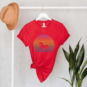 K&ouml;nnte beinhalten: Ein rotes T-Shirt mit einer Einhorn-Silhouette vor einem Sonnenuntergangsdesign. Der Sonnenuntergang hat einen Farbverlauf aus Orange, Gelb und Lila mit horizontalen Streifen. Das Shirt h&auml;ngt an einem wei&szlig;en Kleiderb&uuml;gel.