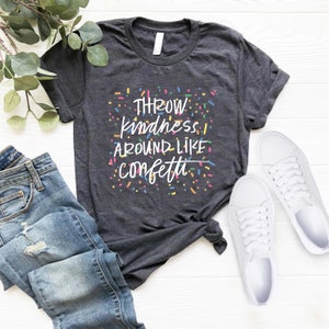 Może przedstawiać: Szary t-shirt z napisem "THROW kindness AROUND LIKE confetti" w kolorze białym z kolorowymi konfetti wok&oacute;ł tekstu.