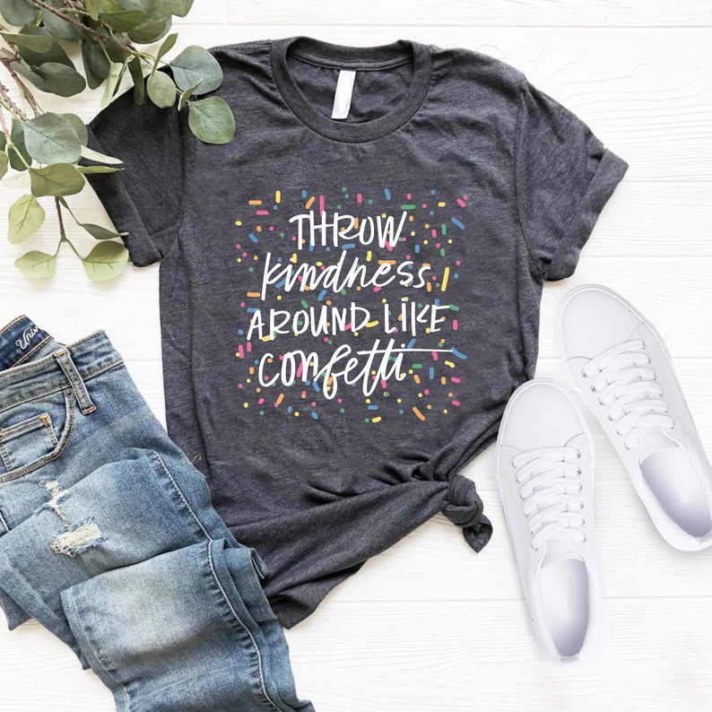 Może przedstawiać: Szary t-shirt z napisem "THROW kindness AROUND LIKE confetti" w kolorze białym z kolorowymi konfetti wok&oacute;ł tekstu.