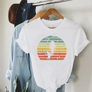 May include: White t-shirt with a vintage-style graphic of a mermaid silhouette in front of a sunset. The graphic has a distressed look with faded colors.