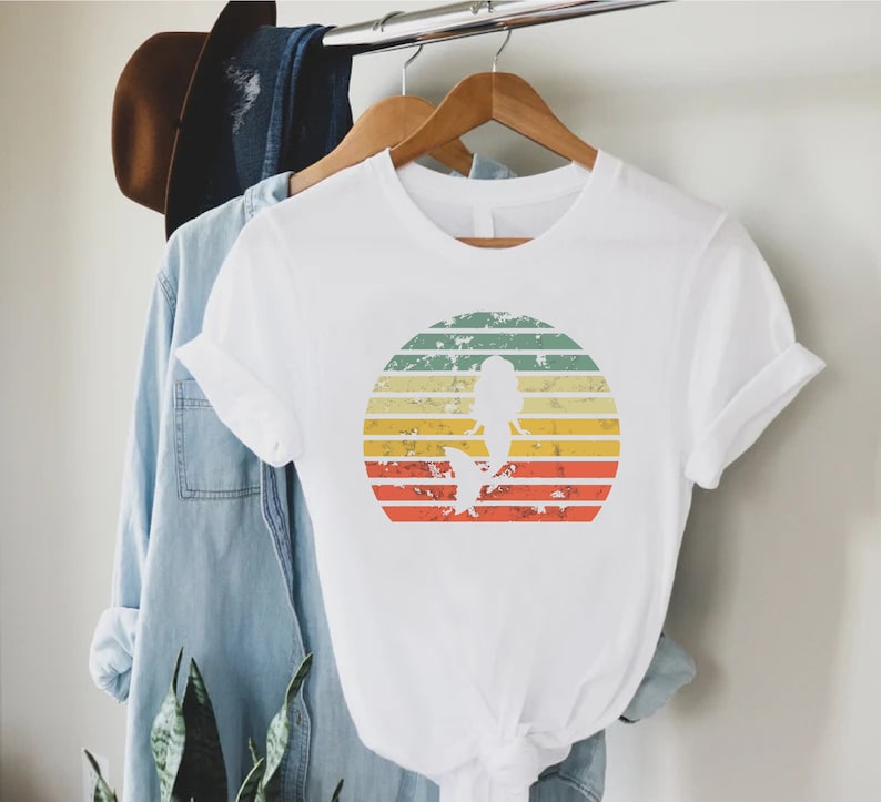 May include: White t-shirt with a vintage-style graphic of a mermaid silhouette in front of a sunset. The graphic has a distressed look with faded colors.