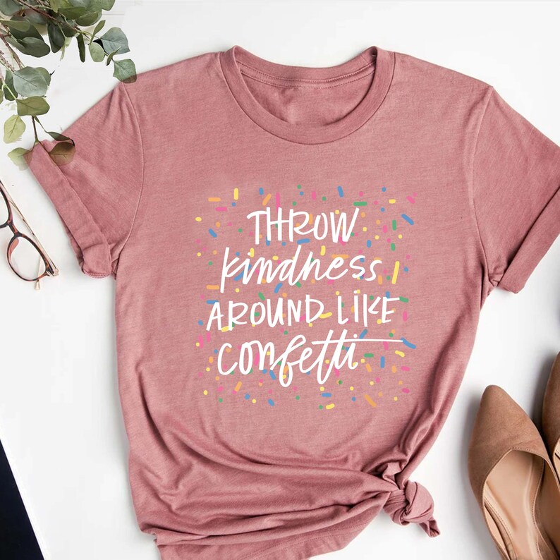 Może przedstawiać: R&oacute;żowy T-shirt z napisem "Throw kindness around like confetti" w białym kolorze. Tekst jest otoczony kolorowymi konfetti.