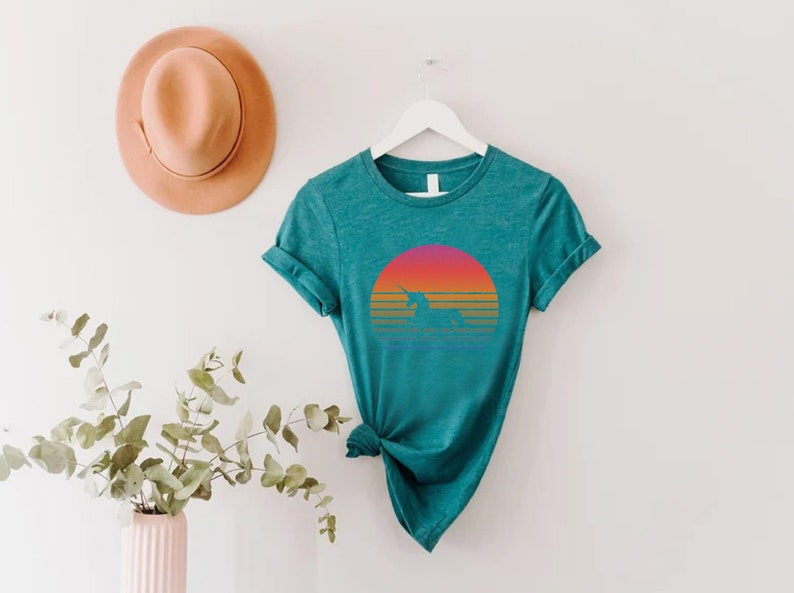 K&ouml;nnte beinhalten: T&uuml;rkisfarbenes T-Shirt mit einer Einhorn-Silhouette vor einem Sonnenuntergangsdesign. Der Sonnenuntergang verl&auml;uft von Pink zu Orange mit horizontalen Linien. Das Shirt h&auml;ngt an einem wei&szlig;en Kleiderb&uuml;gel, mit einem pfirsichfarbenen Hut im Hintergrund.