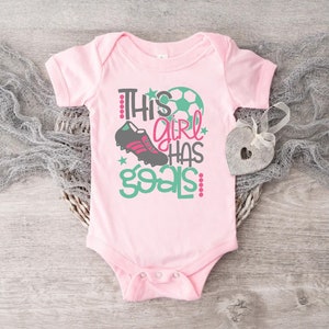 May include: Pink baby onesie with a green and white soccer ball, a black and pink soccer cleat, and the text "This girl has goals" in pink and green.