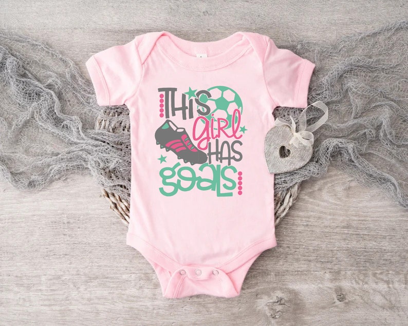 May include: Pink baby onesie with a green and white soccer ball, a black and pink soccer cleat, and the text "This girl has goals" in pink and green.