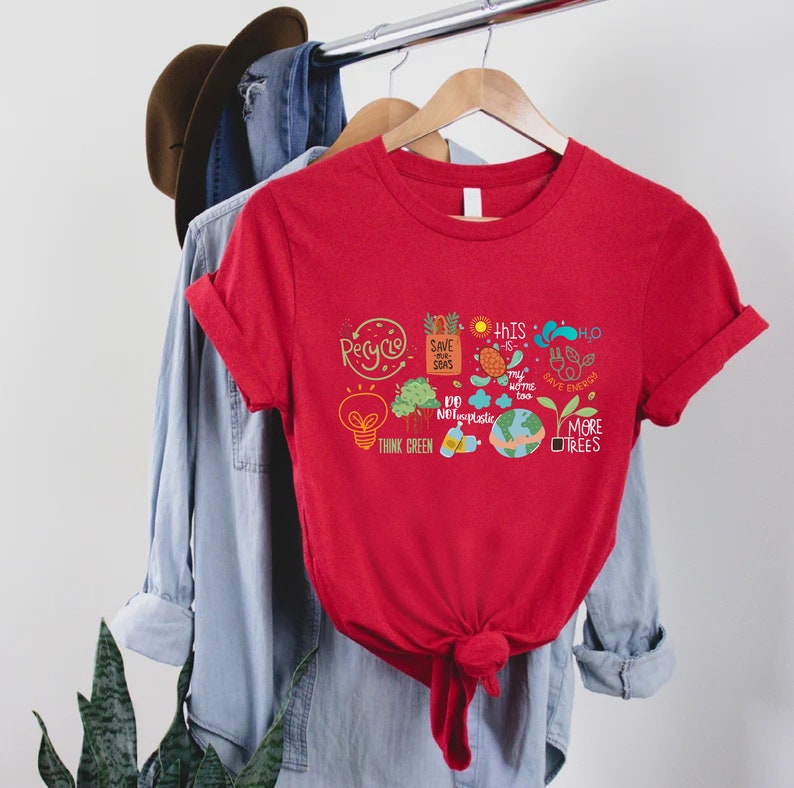 Puede incluir: Camiseta roja con un dise&ntilde;o gr&aacute;fico que presenta una bombilla, &aacute;rboles, un globo terr&aacute;queo y las palabras "Recicla", "Salva nuestros mares", "Piensa en verde", "Este tambi&eacute;n es mi hogar", "No uses pl&aacute;stico", "H2O Ahorra energ&iacute;a" y "M&aacute;s &aacute;rboles".