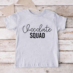 Peut inclure: Un t-shirt gris clair avec le texte "Chocolate Squad" en lettres noires. Le texte est en &eacute;criture cursive avec le mot "Squad" en gras, en caract&egrave;res d'imprimerie.