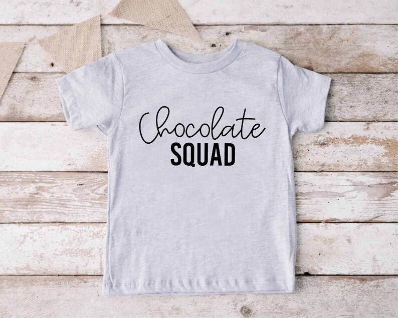 Peut inclure: Un t-shirt gris clair avec le texte "Chocolate Squad" en lettres noires. Le texte est en &eacute;criture cursive avec le mot "Squad" en gras, en caract&egrave;res d'imprimerie.