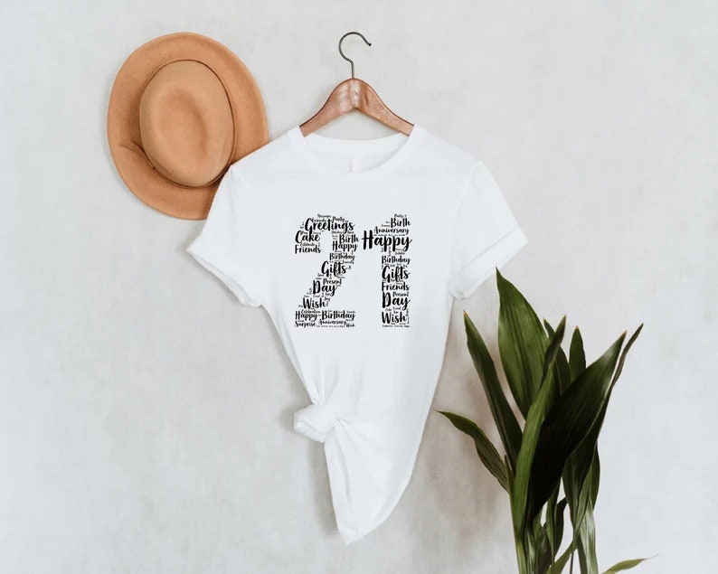 May include: White t-shirt with a black graphic design of the number 21 made up of words related to birthdays, such as "Happy", "Birthday", "Cake", "Gifts", and "Friends".