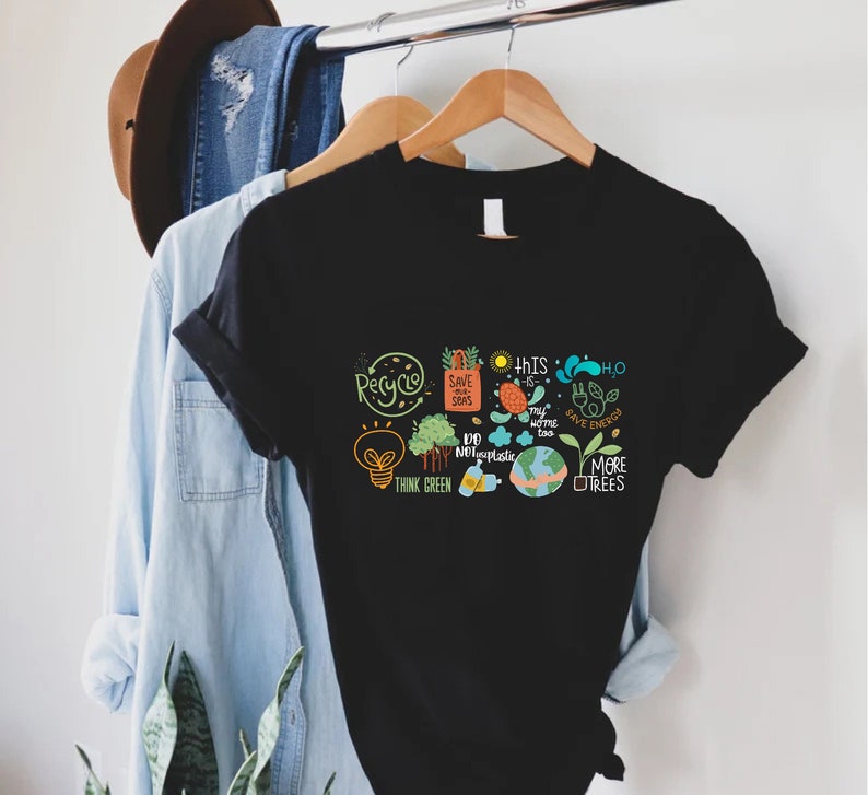 Puede incluir: Camiseta negra con un dise&ntilde;o gr&aacute;fico de varios lemas e ilustraciones ambientales, que incluyen "Reciclar", "Salvar nuestros mares", "Pensar en verde", "No al pl&aacute;stico", "Este tambi&eacute;n es mi hogar", "H2O Ahorrar energ&iacute;a", y "M&aacute;s &aacute;rboles".