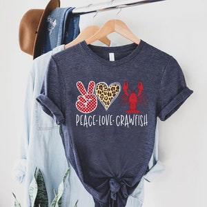 May include: A navy blue t-shirt with a peace sign, a heart with a leopard print, and a red crawfish. The text "Peace Love Crawfish" is printed below the design.