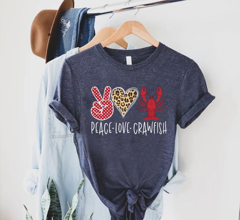 May include: A navy blue t-shirt with a peace sign, a heart with a leopard print, and a red crawfish. The text "Peace Love Crawfish" is printed below the design.