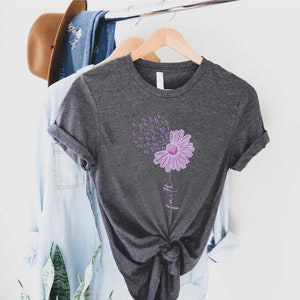 May include: A dark gray t-shirt with a purple flower design and the word "faith" written vertically below the flower. The flower has purple petals and a yellow center. The flower is surrounded by small purple butterflies.