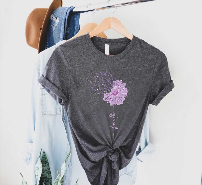 May include: A dark gray t-shirt with a purple flower design and the word "faith" written vertically below the flower. The flower has purple petals and a yellow center. The flower is surrounded by small purple butterflies.