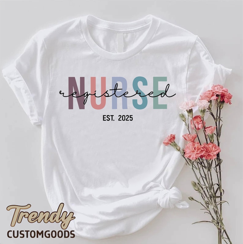 K&ouml;nnte beinhalten: Wei&szlig;es T-Shirt mit dem Text "NURSE registered EST. 2025" in Regenbogenfarben. Der Text ist in einer kursivschrift.