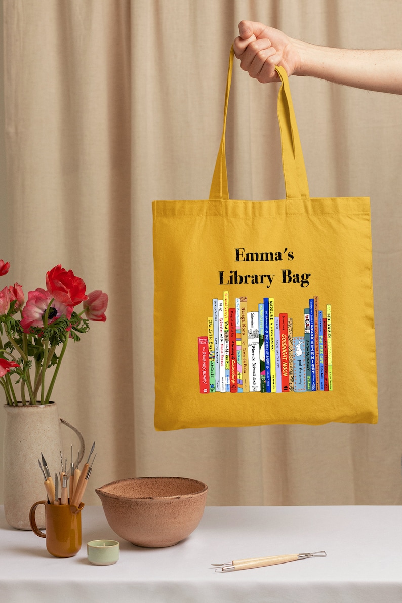 May include: Mustard yellow tote bag with the text "Emma's Library Bag" and a graphic of a stack of colorful books. The bag is held up by a hand, and is made of a sturdy fabric.