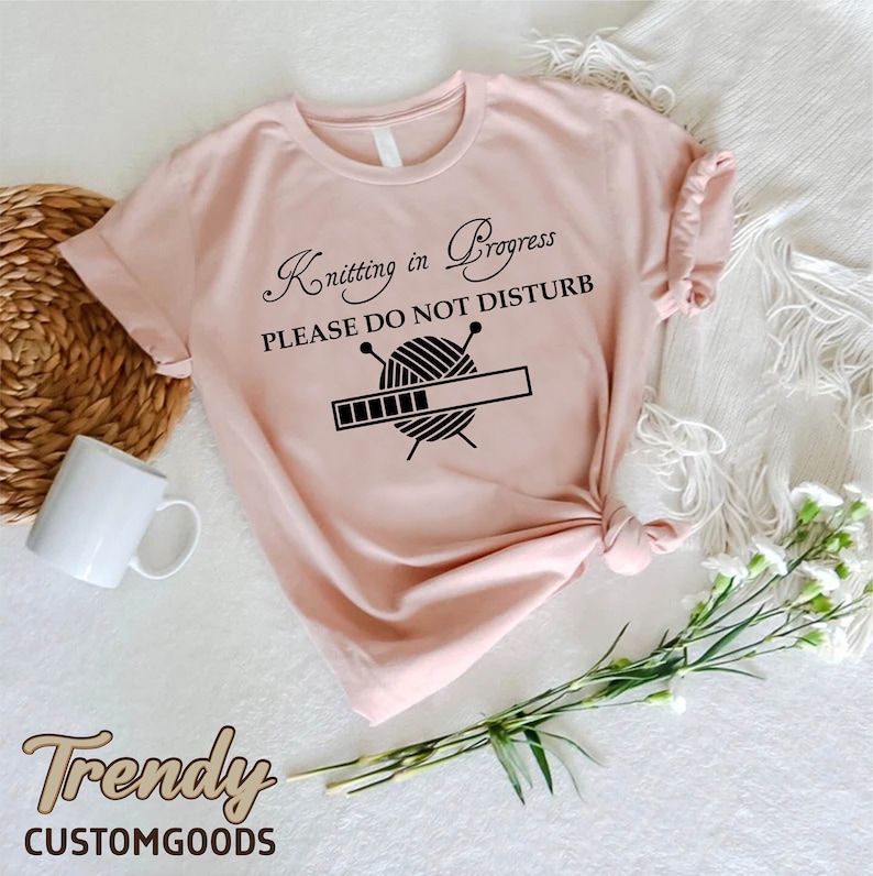 May include: A pink t-shirt with a black graphic that says "Knitting in Progress Please Do Not Disturb" with a yarn ball and knitting needles. The graphic also includes a progress bar.
