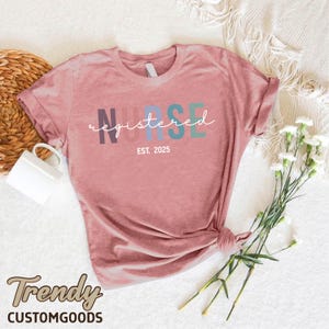 K&ouml;nnte beinhalten: Ein pinkfarbenes T-Shirt mit dem Wort "NURSE" in Regenbogenfarben und dem Text "registered" in wei&szlig;er Schrift darunter. Der Text "EST. 2025" ist unter dem Wort "registered" gedruckt.