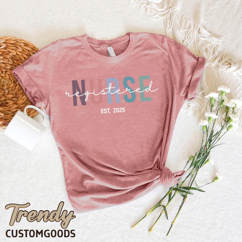 K&ouml;nnte beinhalten: Ein pinkfarbenes T-Shirt mit dem Wort "NURSE" in Regenbogenfarben und dem Text "registered" in wei&szlig;er Schrift darunter. Der Text "EST. 2025" ist unter dem Wort "registered" gedruckt.