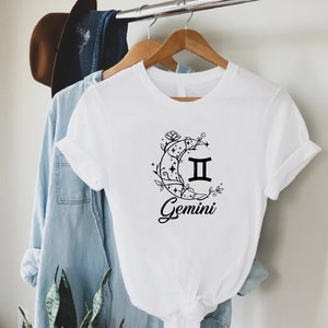 Puede incluir: Camiseta blanca con un gr&aacute;fico negro del signo zodiacal G&eacute;minis dentro de una luna creciente con detalles florales. El texto "Gemini" est&aacute; debajo del gr&aacute;fico.