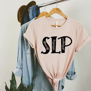 Puede incluir: Una camiseta rosa con las letras "SLP" en negro, con las palabras "speech", "language" y "pathologist" escritas verticalmente dentro de las letras.