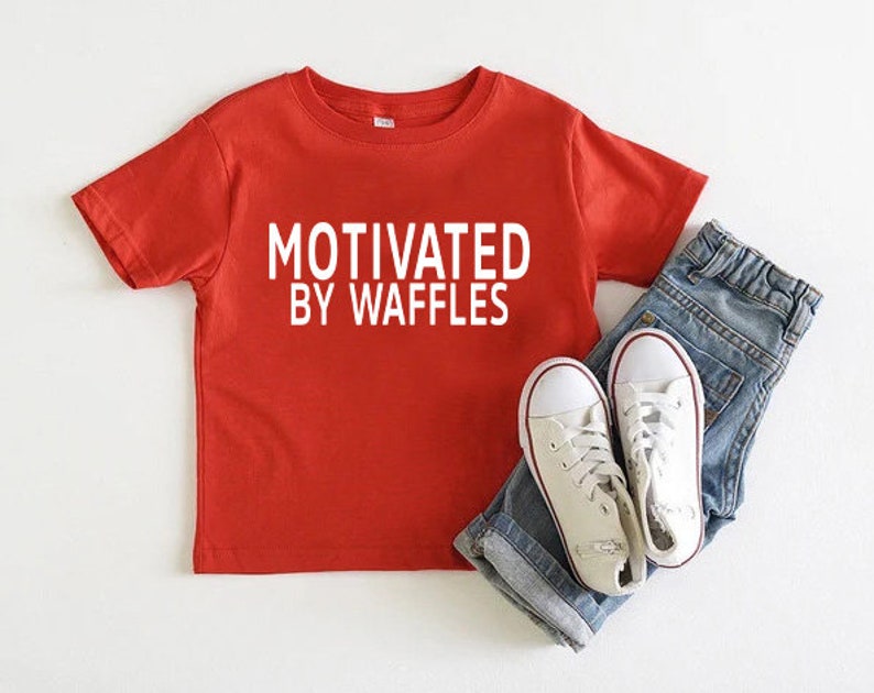 K&ouml;nnte beinhalten: Rotes T-Shirt mit kurzen &Auml;rmeln und wei&szlig;em Text, der "Motivated by Waffles" lautet. Das Shirt wird mit einer blauen Jeans und wei&szlig;en Turnschuhen kombiniert.