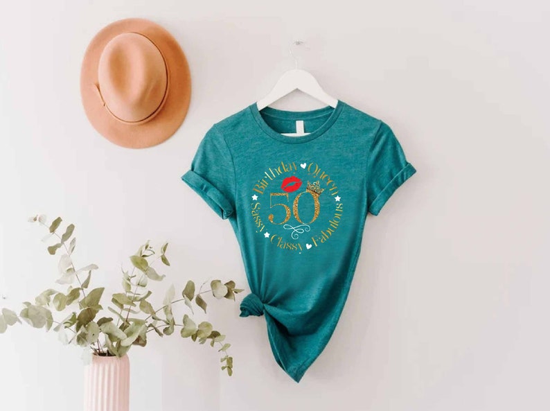 May include: Teal t-shirt with a gold glitter circle design that says "50th Birthday Queen Sassy, Classy, Fabulous". The number 50 is gold glitter and there is a red lip and a gold crown in the circle.