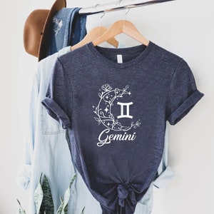 Puede incluir: Una camiseta azul marino con un gr&aacute;fico blanco de una luna creciente con estrellas y enredaderas florales. La luna tiene el s&iacute;mbolo del zod&iacute;aco G&eacute;minis dentro y la palabra "G&eacute;minis" est&aacute; impresa debajo de la luna.