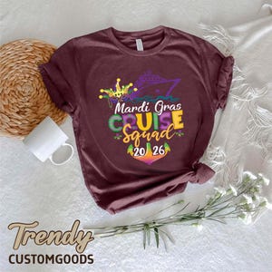 May include: A maroon short-sleeve t-shirt featuring a graphic of a cruise ship, a jester mask, and the text "Mardi Gras CRUISE Squad 20 26" in purple, green, and yellow. The design includes fleur-de-lis accents and a colourful anchor graphic.