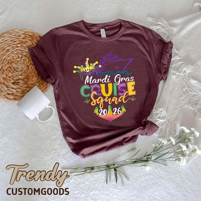 May include: A maroon short-sleeve t-shirt featuring a graphic of a cruise ship, a jester mask, and the text "Mardi Gras CRUISE Squad 20 26" in purple, green, and yellow. The design includes fleur-de-lis accents and a colourful anchor graphic.