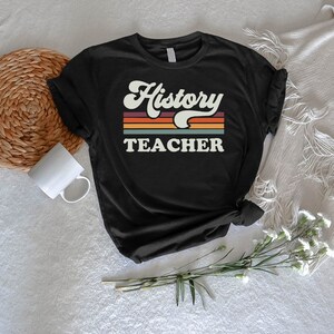 May include: Black t-shirt with a retro-style graphic that says "History Teacher" with colorful stripes behind the text.