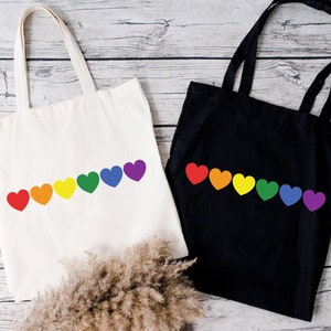May include: Two tote bags with rainbow heart designs. One bag is white and the other is black.