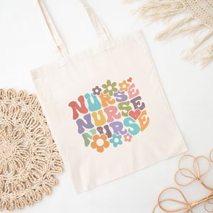 Puede incluir: Una bolsa de lona blanca con un dise&ntilde;o retro colorido que dice "NURSE" en forma de coraz&oacute;n con flores.