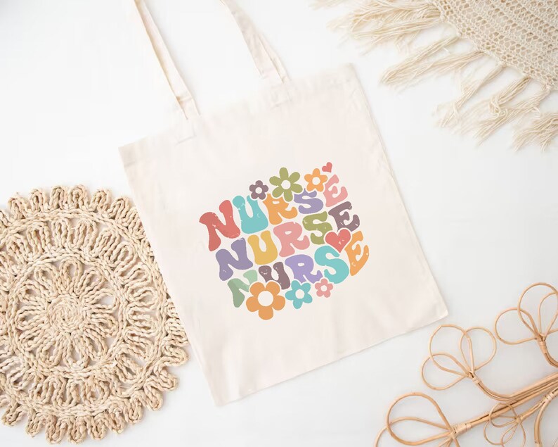 Puede incluir: Una bolsa de lona blanca con un dise&ntilde;o retro colorido que dice "NURSE" en forma de coraz&oacute;n con flores.