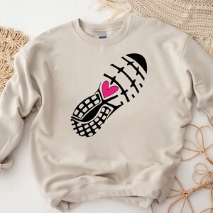 Peut inclure: Un sweat-shirt beige avec un imprim&eacute; noir et blanc d'une chaussure de course avec un c&oelig;ur rose.