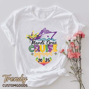 May include: A white t-shirt featuring a graphic of a cruise ship, a jester mask, and the text Mardi Gras CRUISE squad 20 26. The design uses purple, green, and yellow colours with fleur-de-lis accents and an anchor shape around the year 2026.