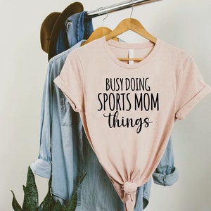 May include: A pink t-shirt with the text "Busy Doing Sports Mom Things" in black lettering.