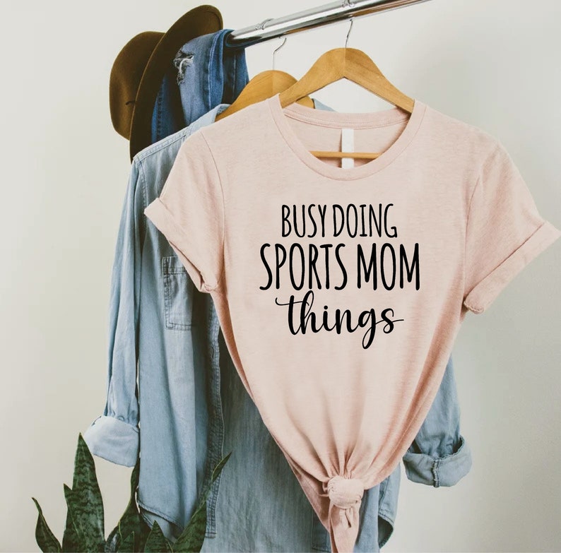 May include: A pink t-shirt with the text "Busy Doing Sports Mom Things" in black lettering.