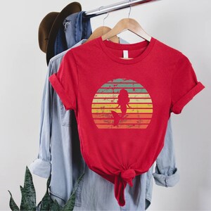 May include: Red t-shirt with a vintage-style sunset graphic featuring a mermaid silhouette. The graphic has a distressed look with faded colors.