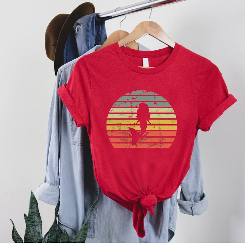 May include: Red t-shirt with a vintage-style sunset graphic featuring a mermaid silhouette. The graphic has a distressed look with faded colors.